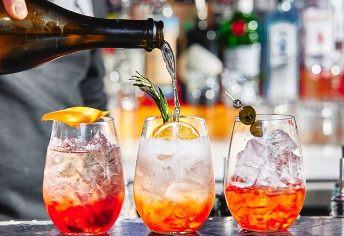 New York venue: Bartender pours a cocktail. Three glasses hold colorful drinks with ice, fruit, and rosemary. Gay travel advice.