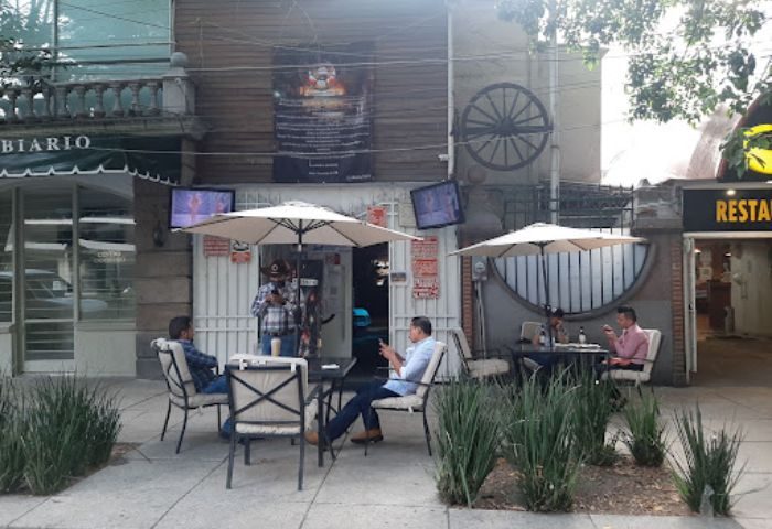 Vaqueros Gay Bar in Mexico: Patrons sit outside at tables under umbrellas. A cowboy-hatted man stands near the entrance.