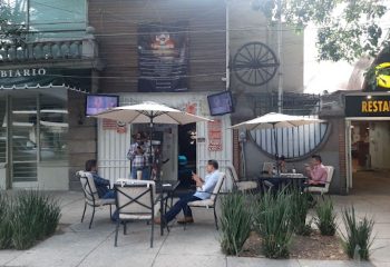 Vaqueros Gay Bar in Mexico: Patrons sit outside at tables under umbrellas. A cowboy-hatted man stands near the entrance.