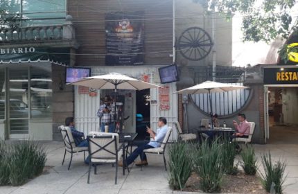 Vaqueros Gay Bar in Mexico: Patrons sit outside at tables under umbrellas. A cowboy-hatted man stands near the entrance.
