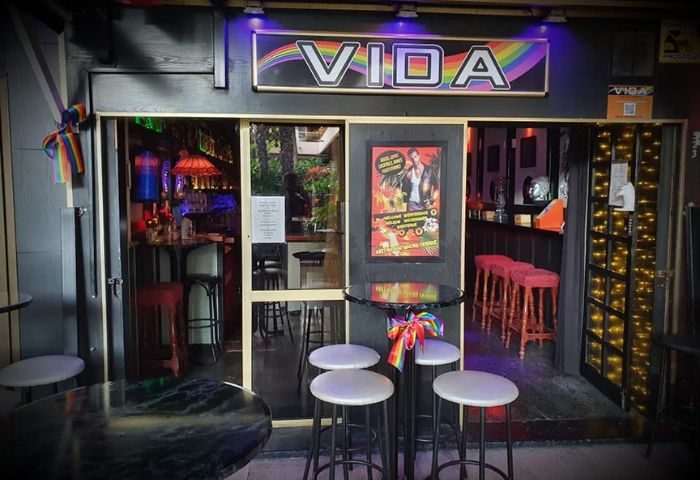 Vida Bar entrance, a gay bar in Torremolinos, Spain. Rainbow flag decor, tables, and bar stools visible. Nightlife scene for gay travelers.