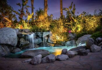 Vista Grande Resort for Gay Men, Palm Springs. Waterfall feature surrounded by lush tropical plants and rocks, illuminated at dusk.