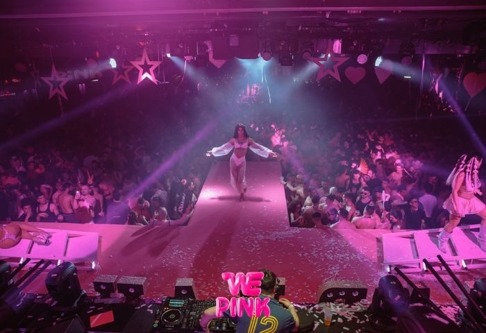 WE Party Madrid gay bar: runway show with a performer on stage in a crowded club. Pink lighting, DJ booth in foreground.