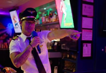 Honolulu's Wang Chung's Karaoke Bar: A person in a pilot costume holds a microphone and points. Gay travel, drag, and drinks in Honolulu.