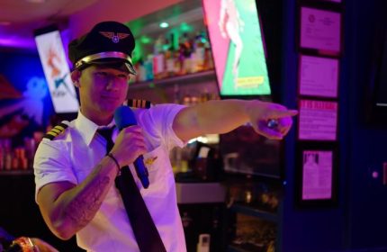Honolulu's Wang Chung's Karaoke Bar: A person in a pilot costume holds a microphone and points. Gay travel, drag, and drinks in Honolulu.