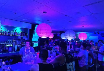 Wang Chung's Karaoke Bar in Honolulu. Gay bar scene with drinks, people, and a bartender under blue and pink lights. Gay travel.