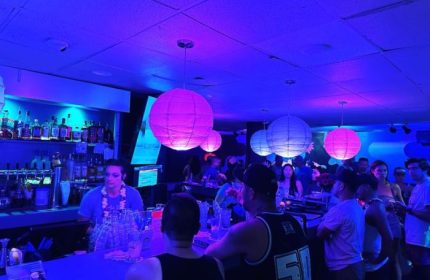 Wang Chung's Karaoke Bar in Honolulu. Gay bar scene with drinks, people, and a bartender under blue and pink lights. Gay travel.