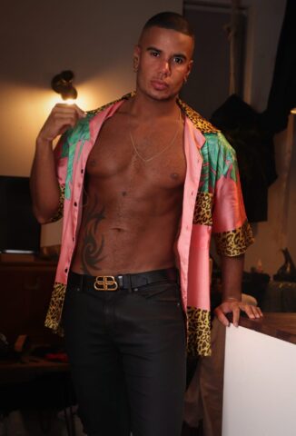Gay travel: Muscular man in unbuttoned tropical shirt, black jeans, and Balenciaga belt. Tattoo on stomach. Stylish, confident.