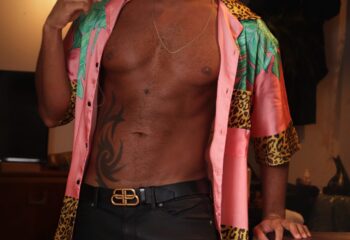 Gay travel: Muscular man in unbuttoned tropical shirt, black jeans, and Balenciaga belt. Tattoo on stomach. Stylish, confident.