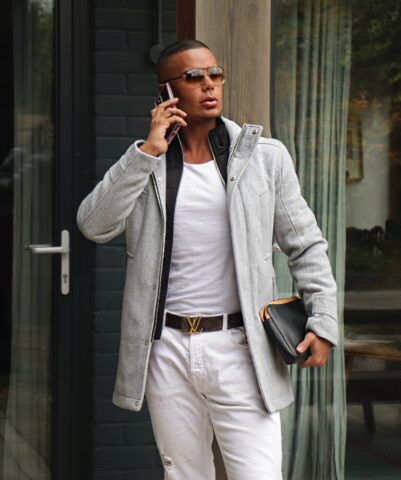 Gay man travel advice: Stylish man in coat, sunglasses, and white jeans talks on his phone, holding a clutch.