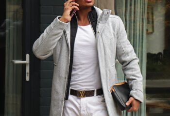 Gay man travel advice: Stylish man in coat, sunglasses, and white jeans talks on his phone, holding a clutch.