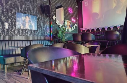 Interior of Why Not Cafe and Bar, Budapest. Stylish seating, dark walls, neon lights, and a large screen create a vibrant, gay-friendly atmosphere.
