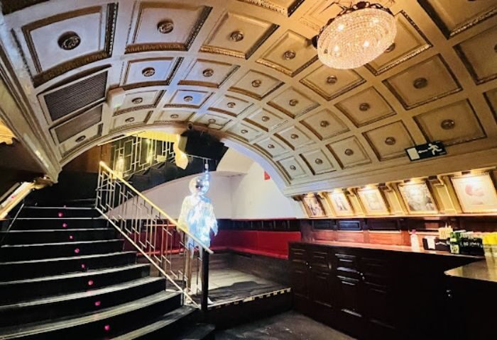 Interior of Why Not? gay bar in Madrid. Stairs lead up to a bar area with red seating, framed pictures, and a decorative ceiling.