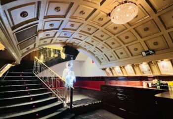 Interior of Why Not? gay bar in Madrid. Stairs lead up to a bar area with red seating, framed pictures, and a decorative ceiling.