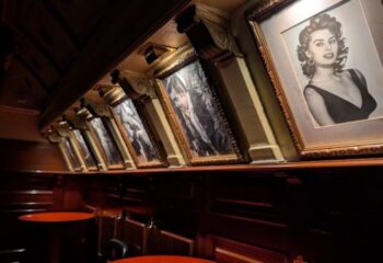 Interior of Why Not? gay bar in Madrid. Framed black and white portraits line the wall above red tables. Gay travel advice.