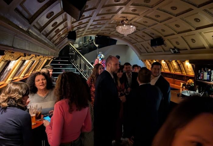 Interior of Why Not? gay bar in Madrid. People mingling under an ornate ceiling with a chandelier, near a bar and staircase.