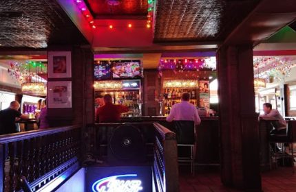 Interior of Woody's gay bar in Toronto. People at the bar, colorful lights, and a Bud Light neon sign. Gay travel advice.