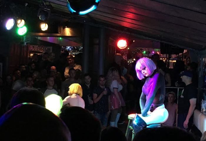 Drag queen performs at Woody's gay bar in Toronto. Nightlife scene with a crowd, stage lights, and performer in a purple wig.