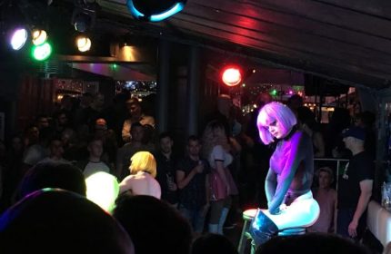 Drag queen performs at Woody's gay bar in Toronto. Nightlife scene with a crowd, stage lights, and performer in a purple wig.