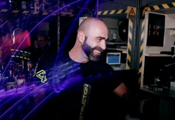 Woof X Lisbon: Smiling bald man at a bar, possibly a bartender. Nightlife scene. Gay travel in Lisbon, Portugal. Nightclub atmosphere.