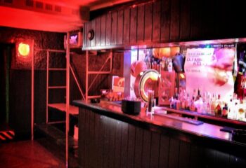 Woof X Lisbon bar interior. Red lighting, wooden bar, liquor bottles, and a DJ poster. Gay travel advice for Lisbon's nightlife scene.