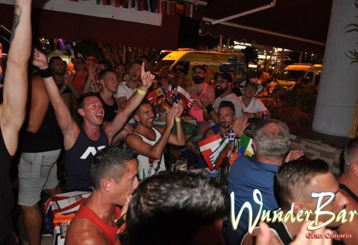 Crowd at WunderBar Gay Bar in Gran Canaria cheering with flags. Lively atmosphere, international flags, and enthusiastic gay men enjoying the nightlife.