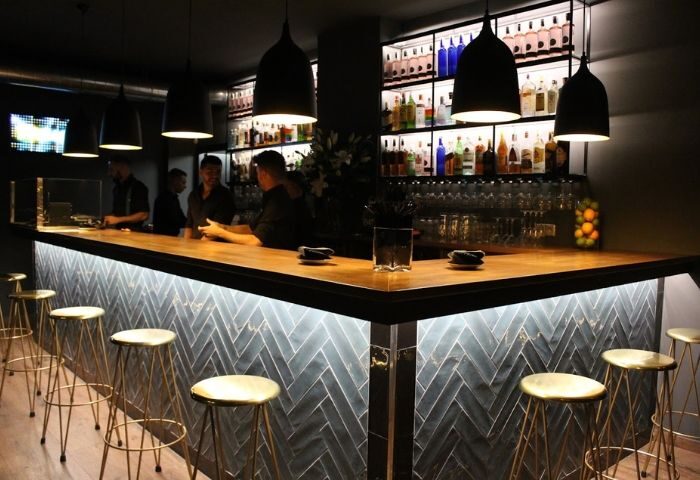 YOUME Gay Bar in Madrid: Interior view of a stylish bar with stools, bartenders, liquor bottles, and pendant lights. Gay travel advice.