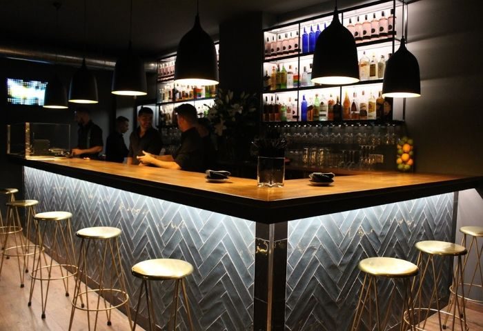 YOUME Gay Bar in Madrid: Interior view of a stylish bar with stools, bartenders, liquor bottles, and pendant lights. Gay travel advice.