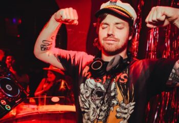 DJ at Yah Yah's Melbourne. Gay nightlife travel. Man with Captain hat flexing, tattoos, and headphones. Red lighting. Gay travel guide.