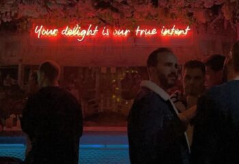 Young cool gay crowd at The Friendly Society, a London gay bar. Red neon sign reads Your delight is our true intent. Nightlife scene.