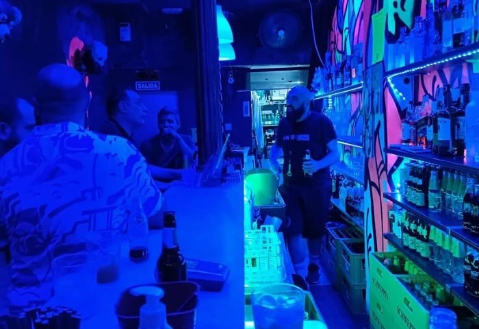 Interior of Zarpa Bear de Copas, a gay bar in Madrid. Bartender with beard, drinks, and shelves of liquor. Blue lighting.