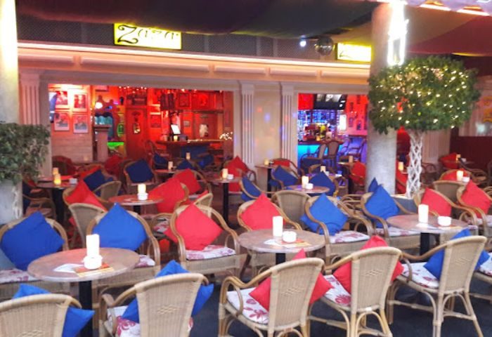 Outdoor seating area at Zaza Bar, a gay bar in Gran Canaria. Wicker chairs with blue and red cushions surround small tables. Interior is visible.