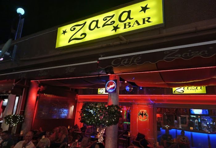 Zaza Bar, a gay bar in Gran Canaria, Spain. Exterior view at night with patrons seated at tables. Red lighting and illuminated signs.