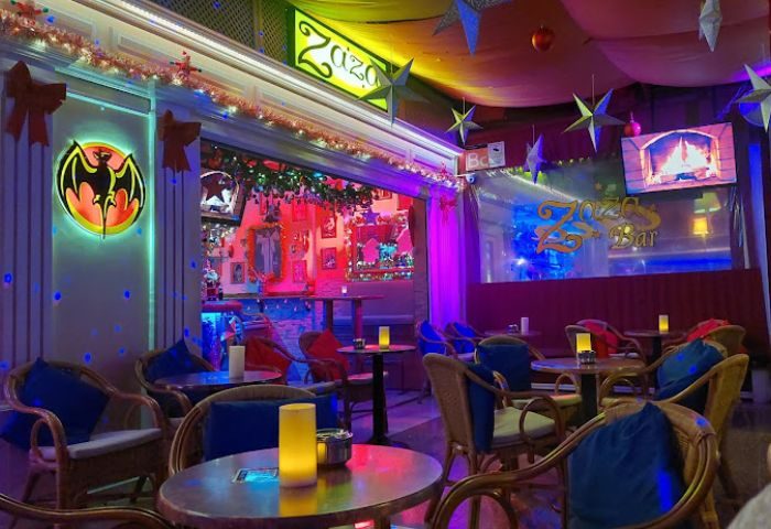 Interior of Zaza Bar, a gay bar in Gran Canaria. Tables, chairs, festive decor, and a Bacardi logo are visible. Nightlife scene.