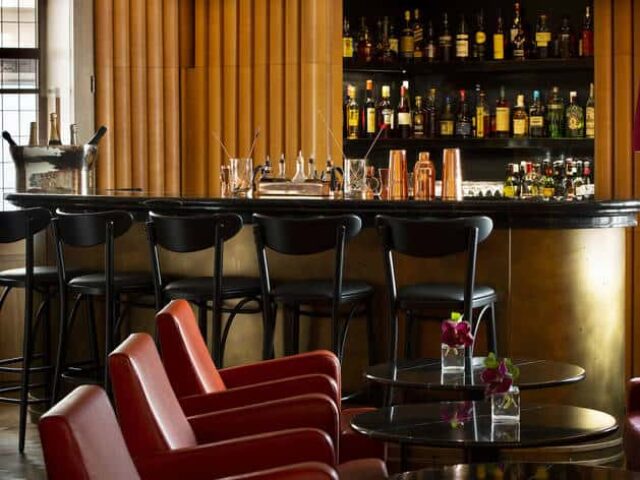 Interior of Ziggy's Gay Bar. Bar stools line the counter, with liquor bottles on shelves. Red armchairs and tables in foreground.
