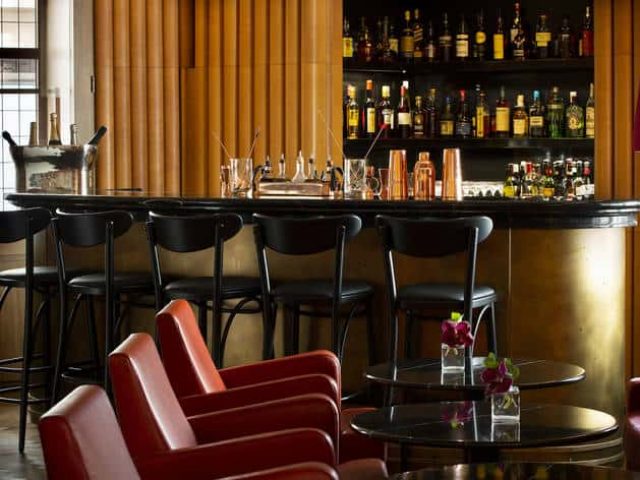 Interior of Ziggy's Gay Bar. Bar stools line the counter, with liquor bottles on shelves. Red armchairs and tables in foreground.
