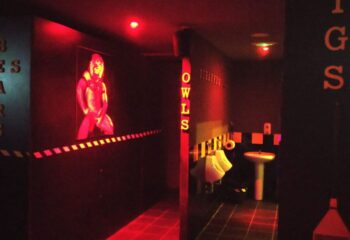Zoo Men's cruising bar in Gran Canaria. Red-lit interior with urinals, sink, and a suggestive image. Gay cruise club atmosphere.