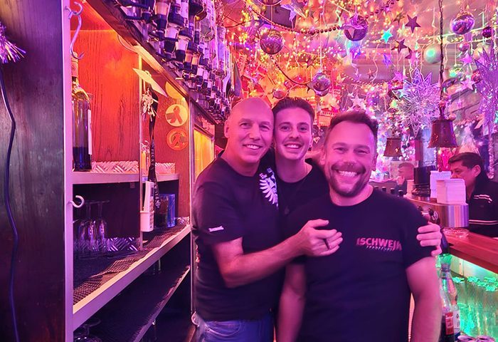 Three smiling men at Zum Schwejk, Frankfurt. Gay bar with festive decor, bottles, and glasses. Gay travel advice.