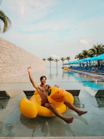 Gay Songkran travel: Man on inflatable duck in pool. Tropical resort with palm trees. Gay travel advice, fun vacation, a5732f99-eb96.