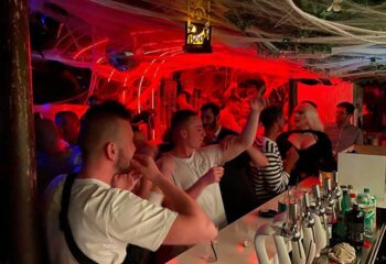 Inside Banana Cafe, a gay bar, patrons celebrate Halloween with red lights, spiderwebs, and a Boo sign. Drinks line the bar.
