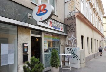 Exterior of Café Rifugio in Vienna, a gay-friendly café. Features signage, street view, and a rainbow flag reflection in the window.