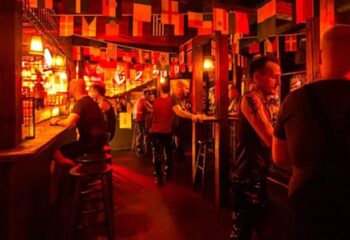 Inside EAGLE Bar Vienna, a popular gay venue. Men in leather attire mingle under a ceiling decorated with international flags. Red lighting creates a vibrant atmosphere.