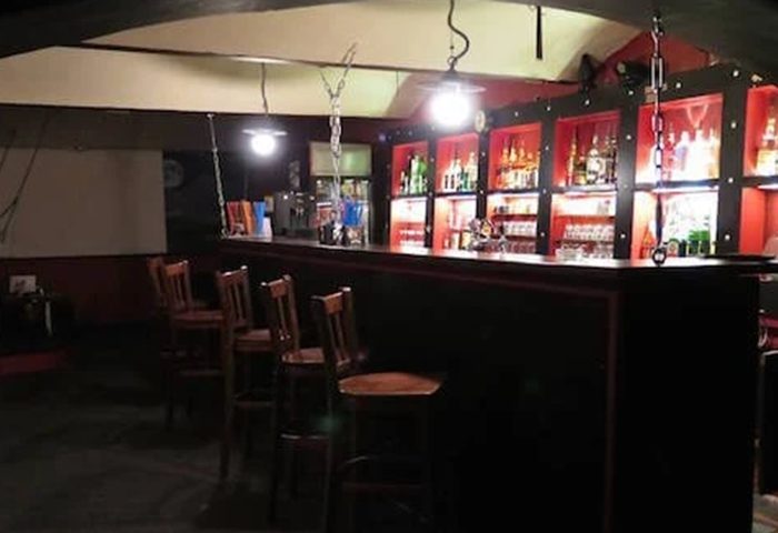 Interior of Factory Club Prague, a gay bar. Dark bar counter with wooden chairs, shelves of liquor bottles, and industrial-style lighting.