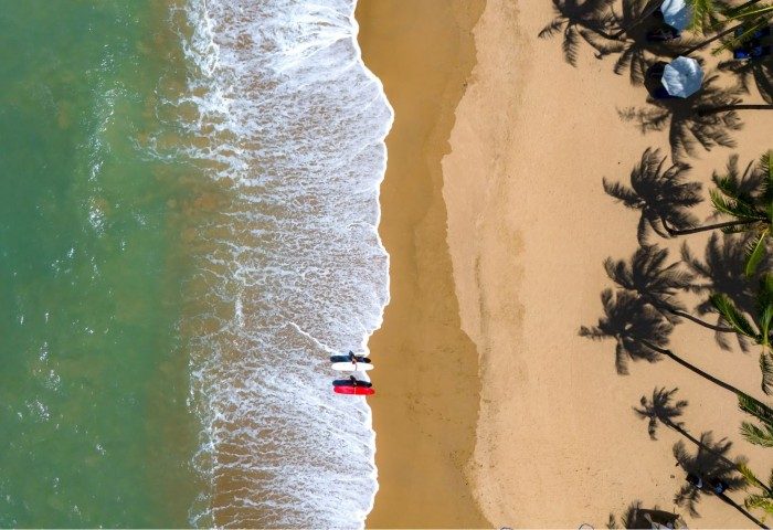 Aerial view of JW Marriott Khao Lak Resort's beach. Palm trees, surfboards, and ocean waves create a surfer paradise. Gay travel.
