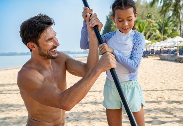 JW Marriott Khao Lak Resort & Spa watersports: A gay man helps a child with paddleboarding on the beach, Thailand. Classic family fun.
