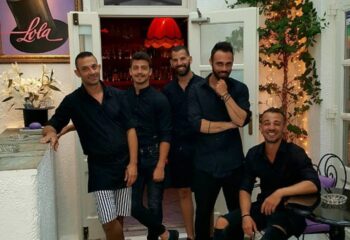 Staff at Lola Bar in Mykonos. Five handsome gay men in black shirts pose at this popular gay travel destination. Nightlife scene.