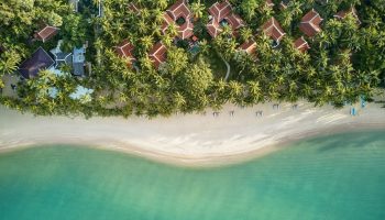 Aerial view of Santiburi Beach, Koh Samui. White sand, turquoise water, palm trees, and resort buildings. Perfect for gay men's travel advice.