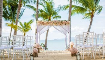 Santiburi Koh Samui wedding arch. Beach wedding setup with white chairs, floral arch, palm trees, and ocean view. Gay travel destination.
