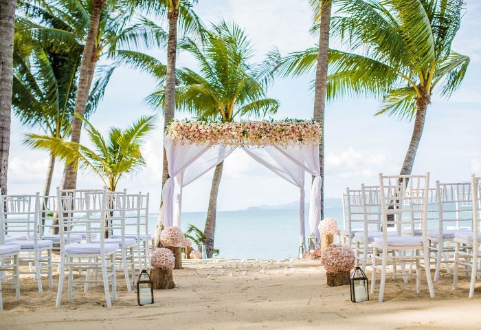 Santiburi Koh Samui wedding arch. Beach wedding setup with white chairs, floral arch, palm trees, and ocean view. Gay travel destination.