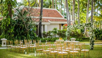 Santiburi Koh Samui wedding seating. Gold chairs face a floral archway on green grass. Palm trees and a building are in the background.
